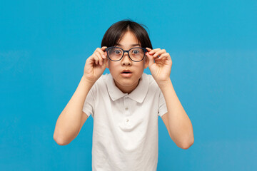 shocked asian boy schoolboy of twelve years old looks into glasses and is surprised on blue isolated background