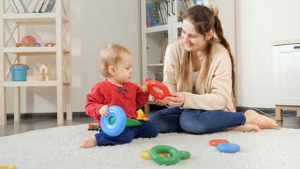 Happy mother with child building toy tower on soft carpet at home. Baby development, child playing games, education and learning.