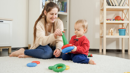 Happy babyboy with mother playing with colorful toys on carpet in living room