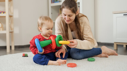 Young mother teaching her little baby son building colorful toy pyramid at home. Baby development, child playing games, education and learning.