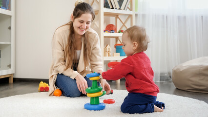Happy smiling mother looking at her little baby son assembling toy pyramid or tower. Baby development, child playing games, education and learning.