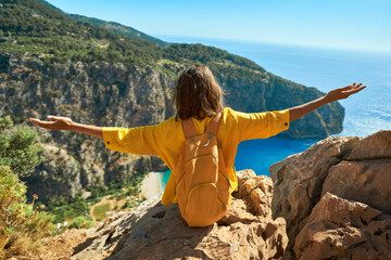 Young woman tourist sitting on cliff with orange backpack freedom opening arms, gazing at the incredible panoramic view of Butterfly Valley Beach