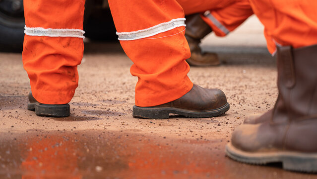 The worker in orange coverall and leather safety shoe is standing on the dirt gorund at construction worksite. Industrial worker in action scene. selective focus.