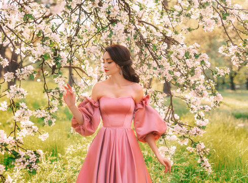 Fantasy girl princess beauty smiling face hand touching white flowers apple tree spring nature green grass forest. Woman queen brunette hair long pink dress puffed sleeves vintage old style art photo