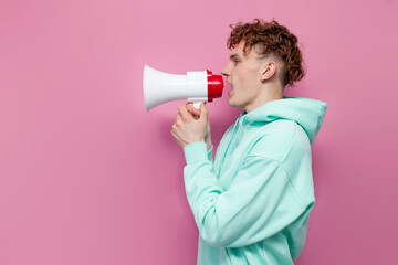 young curly guy in mint hoodie announces and speaks into megaphone on pink isolated background, man...