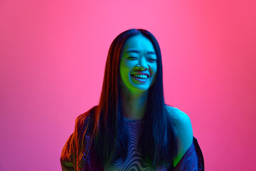 Portrait of happy, smiling, asian, young girl cheerfully posing against pink studio background in neon light. Happiness, success. Concept of emotions, facial expression, youth, lifestyle, sales, ad