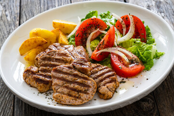 Grilled pork loin steaks with fresh vegetable salad and fried potatoes on wooden table
