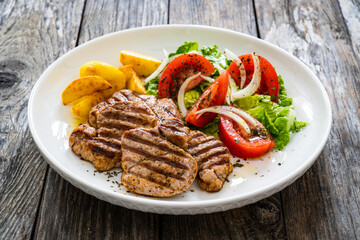 Grilled pork loin steaks with fresh vegetable salad and fried potatoes on wooden table

