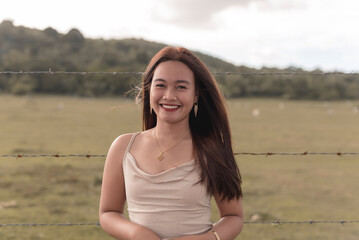 A gorgeous lady in a pink fitted dress smiling widely while standing near the pasture. Sky, trees and a pasture are in the background.