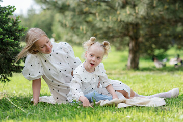 Blond young mother with disabled daughter sitting ot the green grass in the park. Child cerebral palsy. Disability. Inclusion. Means of rehabilitation. Orthosis. Family with disabled kid.