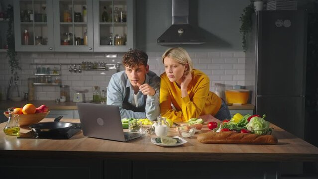 Young romantic couple standing at kitchen island, learning online cooking from laptop computer