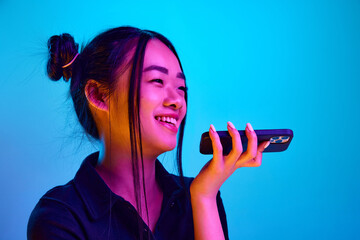 Portrait of young, happy, korean girl smiling and recording voice message on phone against blue...