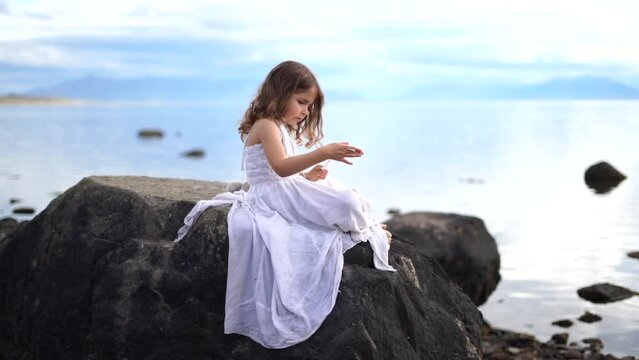 Little girl with white long dress and long hair sitting on big black rock playing with feather on a beach. . High quality FullHD footage