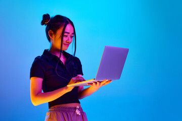 Portrait of young korean girl, student standing with laptop against blue studio background in neon...