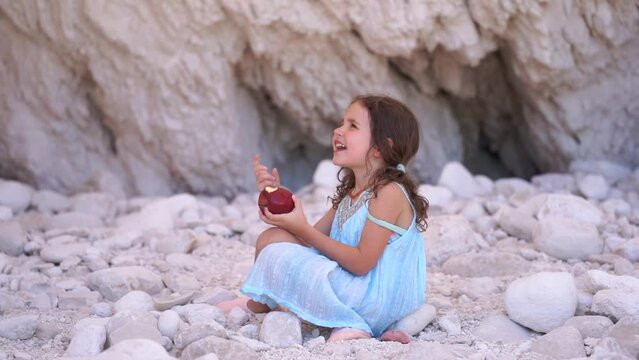 Little girl in blue summer dress eating red apple and looking at something, smiling. in rock beach in Greece. High quality FullHD footage