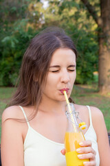 Attractive young girl is drinking sour juice from the bottle through the straw on the bench in the park