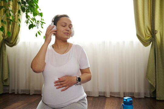 Multi ethnic pregnant woman listens to soothing music on wireless headphones, training yoga in hero pose, gently caressing her belly thinking about her baby and enjoying her future life and pregnancy