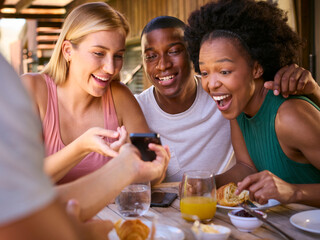 Group Of Smiling Multi-Cultural Friends Eating Breakfast Outdoors At Home Looking At Mobile Phone