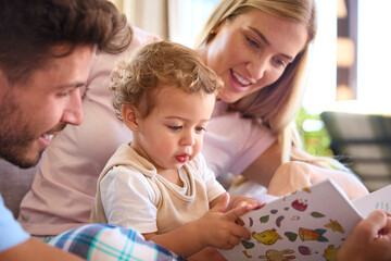 Family Sitting On Sofa At Home With Parents Reading Book With Young Son