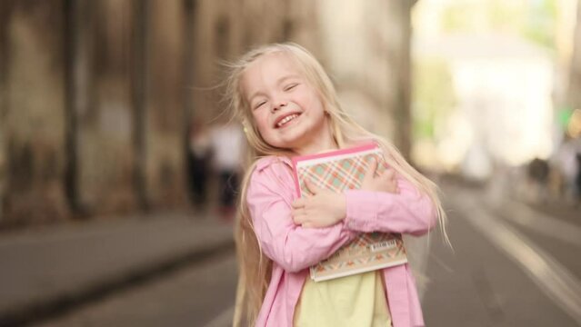 Portrait of overjoyed cute little blond girl with backpack hold notebooks and get back to home after study lessons from school jumping at the urban city street alone Happy childhood concept