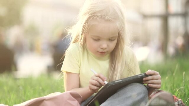 Charming little girl child drawing sketch from nature using tablet at park Pretty kid with long blond hair painting by stylus at screen of digital pad outdoors 