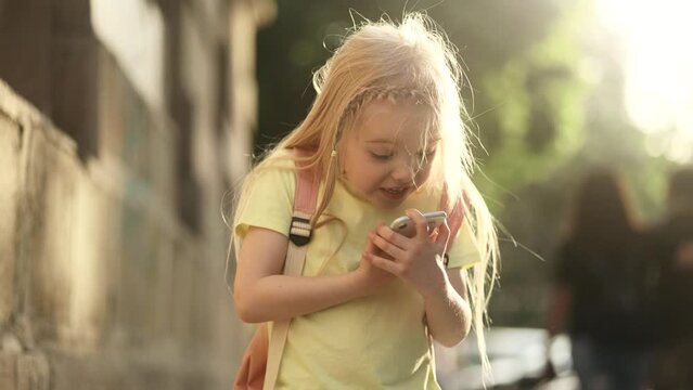 Shocked cute little girl with backpack opening mouth looking at smartphone screen after lessons at city street Adorable kid showing shock or wow emotion by unexpected good news and dancing outdoors