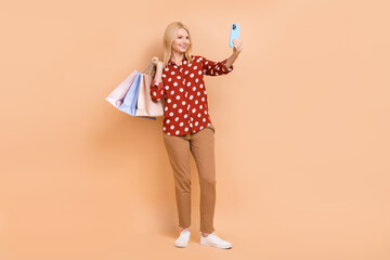 Full length photo of nice positive aged lady hold store bags smart phone make slefie isolated on beige color background