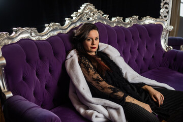 Brunette with red lipstick in a white fur coat on a purple sofa