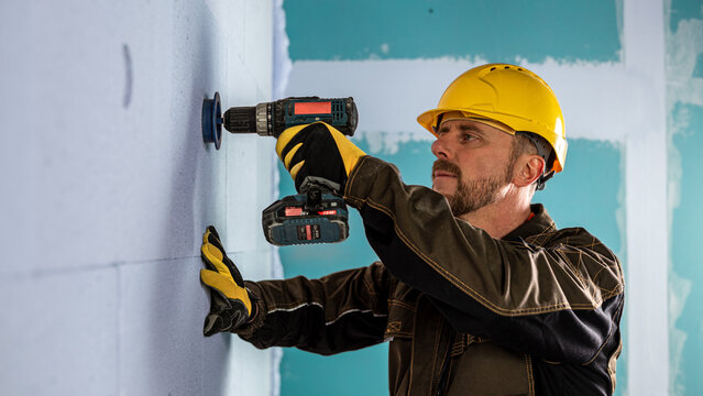 A man dressed work clothes and a helmet working with a cordless drill at milling hole to mineral insulation board.