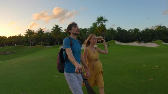A slow-motion stock video captures a pregnant woman and her husband walking hand in hand on a green field during a beautiful sunset. They share a joyful moment, capturing their love and anticipation