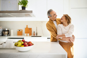 Romantic married senior spouses embracing in kitchen, loving old couple enjoying time together at...