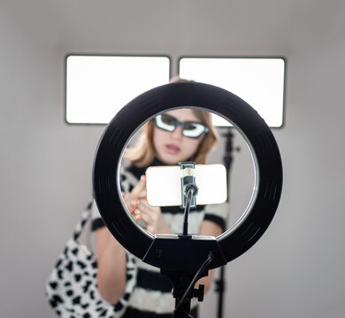 Influencer young woman recording video on smartphone and lighting herself with ring lamp. Broadcast communicates with followers in social networks.