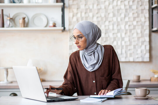 Young muslim woman in hijab freelancer working from home in kitchen. He sits focused on the laptop and makes notes in the notebook.