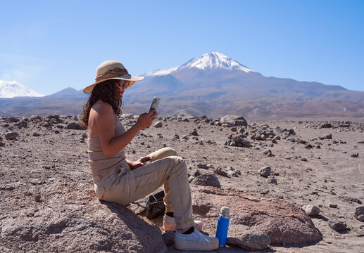 mid adult brunette hiker woman sitting on a rock drinking coffee and taking pictures with her cell phone in San Pedro de Atacama