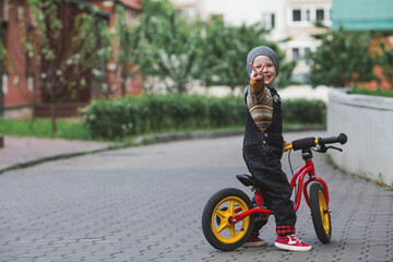 A cheerful little boy rides a bicycle outdoors. A happy child walks in the spring park. The kid is dressed in a fashionable sweater and denim overalls.