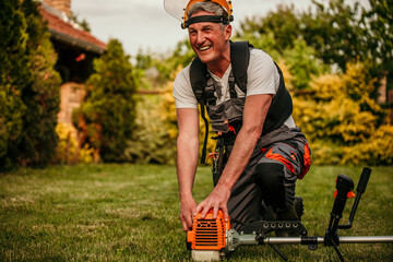 Senior man with brush cutter on the lawn against the background of bushes in workwear and goggles