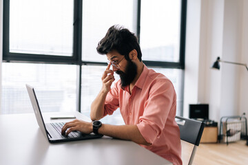 Thoughtful Indian programmer male in casual clothes working on laptop sitting at table in light coworking office on background of window. Bearded business man in glasses looking at computer screen.