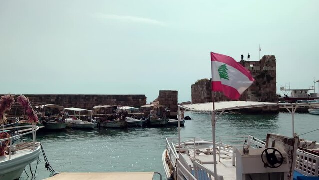 Boats rocking on waves in ancient Port of Byblos. Lebanese flag is waving on a flagpole opposite ruins of port. Byblos ancient city, Jbeil, Lebanon