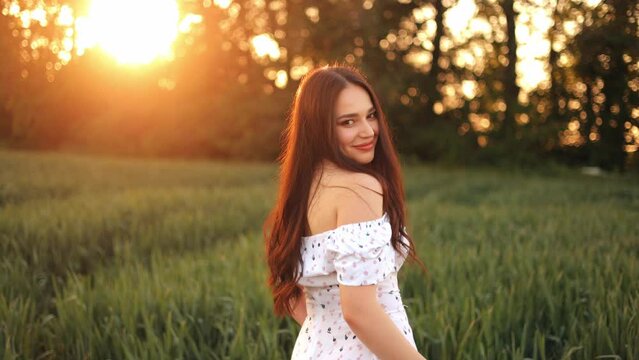 A young girl in a summer dress walks in slow motion through a green field of wheat. Beautiful carefree woman enjoying nature and sunlight in wheat field at sunset