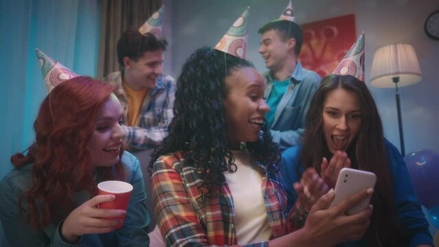 An African American girl shows the phone to her friends, they are emotionally looking at photos, videos or talking on a video call. A group of friends in party hats at a house party. Slow motion.