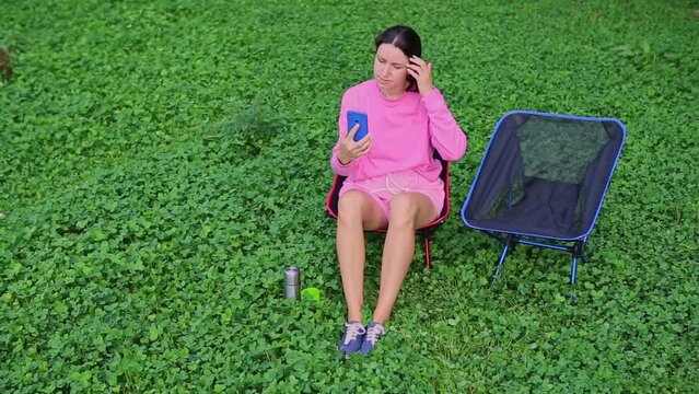 A beautiful brunette girl in a pink sweater looks into the phone camera and straightens her hair and presses the button for selfie in nature. A tanned young woman sits in a camping chair in the park