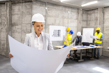 Female structural engineer analyzing building plans and blueprint at construction site while architects brainstorming in background.