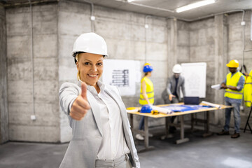 Portrait of successful female architect in business suit and hard hat holding thumbs up at construction site.