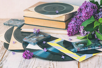 Vinyl records, cassette tapes and stack of books with purple lilac flowers near them.