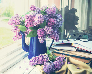 A blue vase of lilacs sits on a window sill next to a stack of books