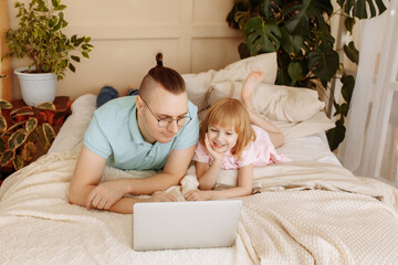 Obraz na płótnie Canvas Handsome young man at home with his little cute daughter are lying on the bed with looking into the laptop. Happy Fathers Day.