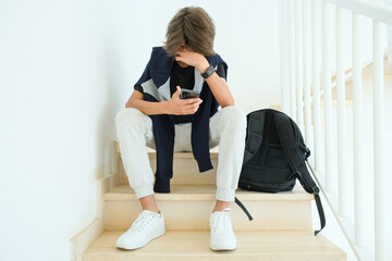 Fototapeta Teenage boy using smartphone. Unhappy, sad, frustrated teenage boy sitting alone on stairs. Learning difficulties, gaming addiction, emotion, psychological problems in adolescence concept obraz