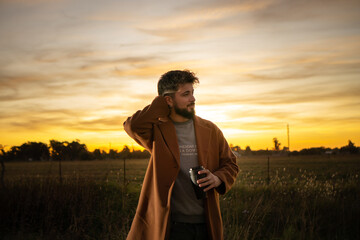 young man drinking coffee in the field with a sunset in the background