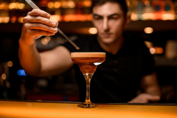 Bartender holds tweezers with coffee bean and decorates by it glass with espresso martini cocktail