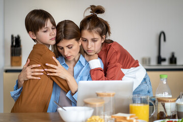 Portrait of sad mother hugging with kids son and daughter while sitting in front of laptop computer in kitchen, children embracing and supporting tired single mom, family having financial problems